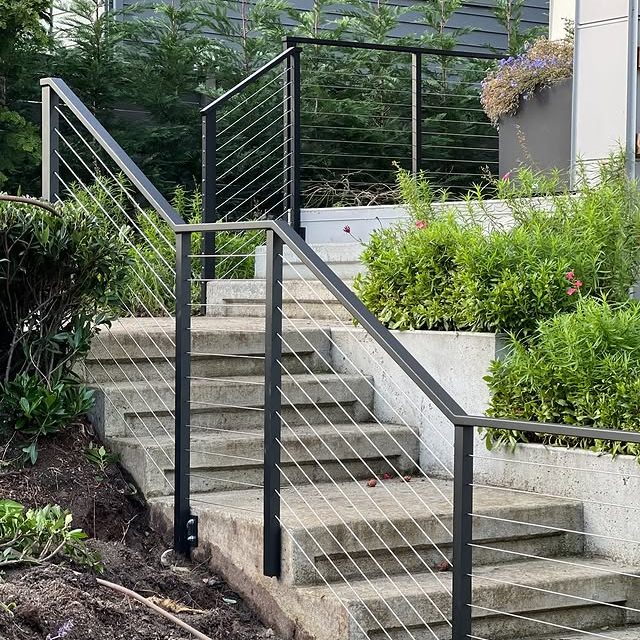 Black metal stair railing with stainless steel cable infill installed on an outdoor concrete staircase with surrounding greenery.